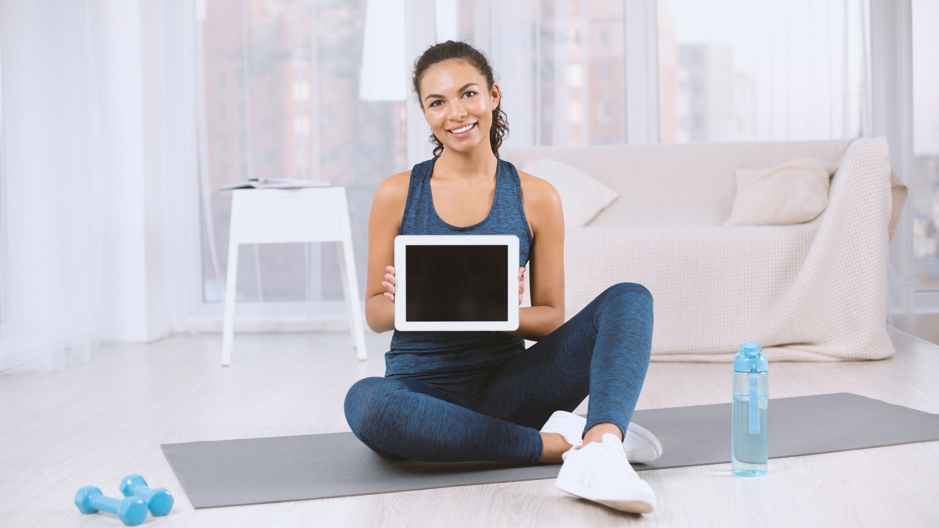 Happy Latin woman showing tablet computer with empty screen on yoga mat at home, mockup for design